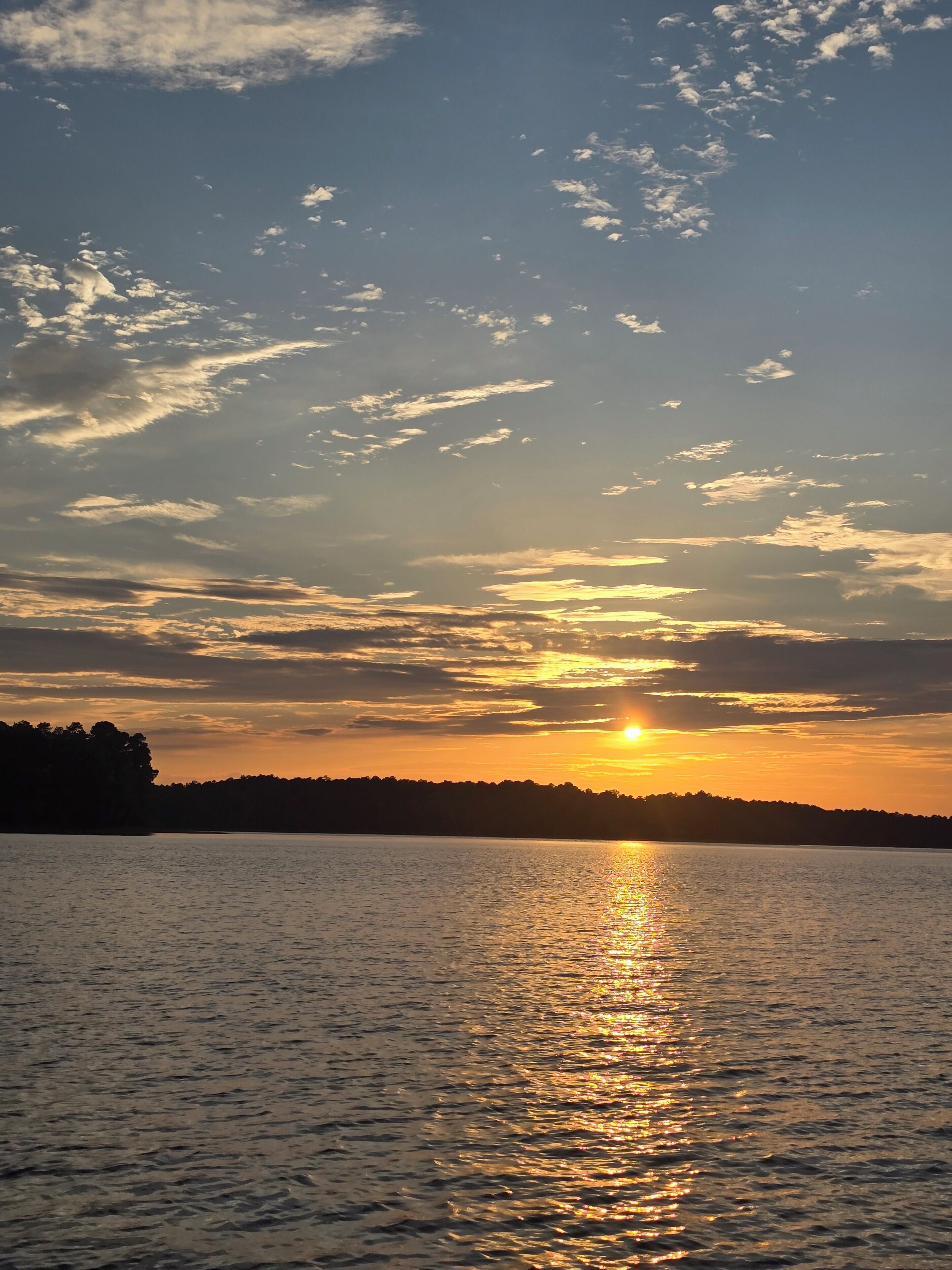 Ein Sonnenuntergang über ruhigem Wasser, im Hintergrund die Silhouette von Bäumen.