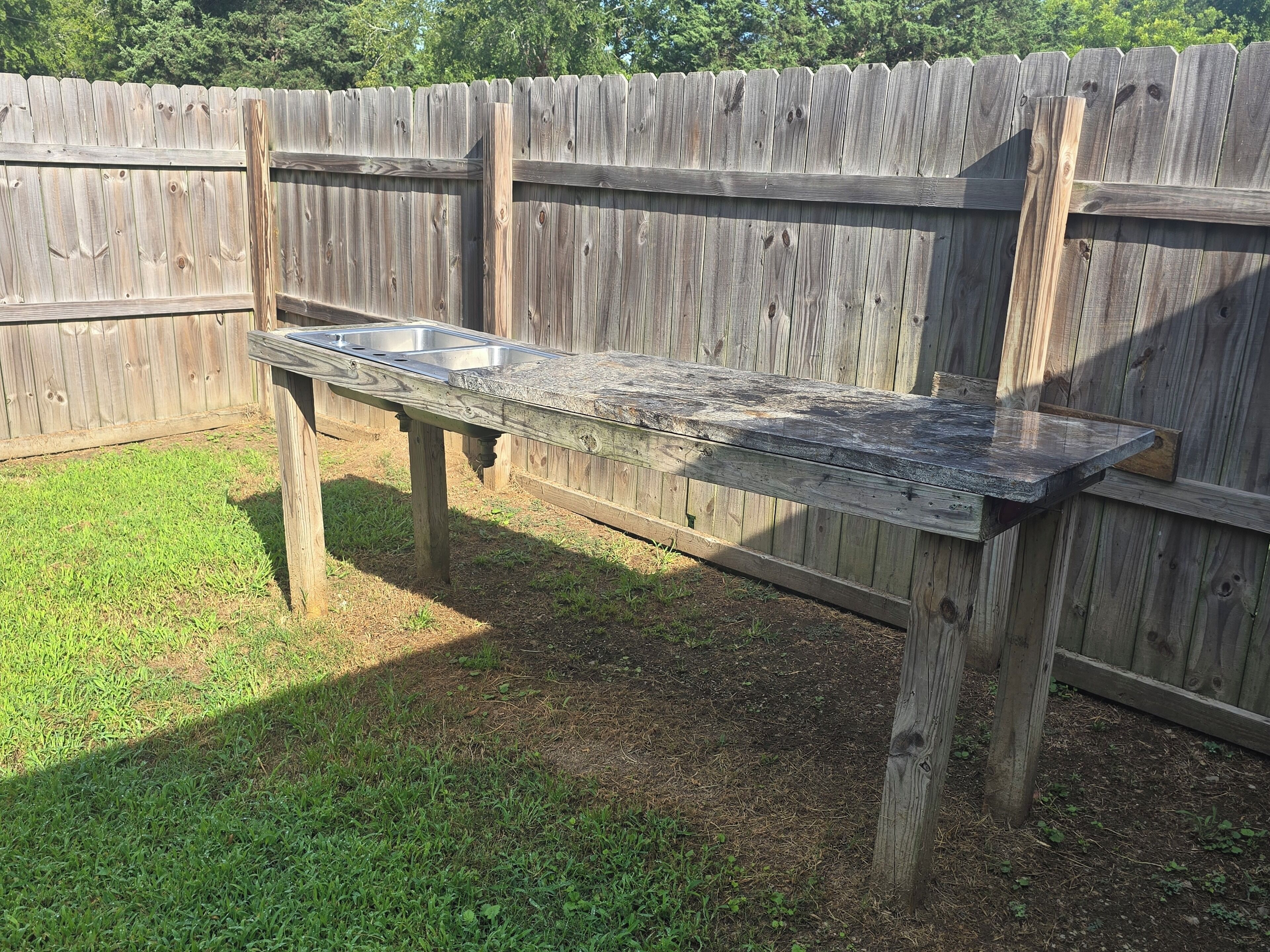 A wooden picnic table with metal containers, set against a wooden fence in a grassy area.