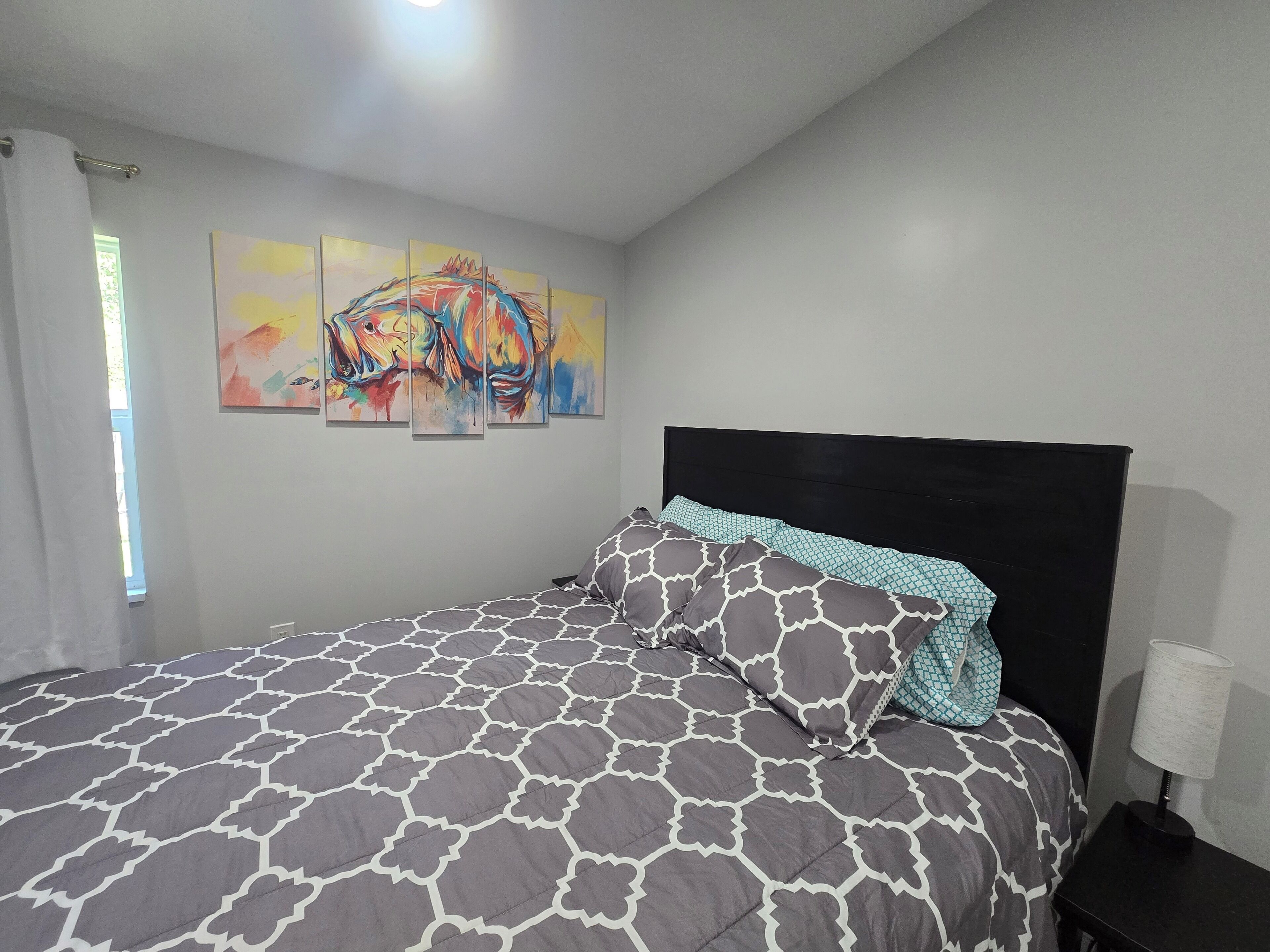 Une chambre à coucher avec un lit, une table de chevet, une lampe et un mur orné d’un tableau.
