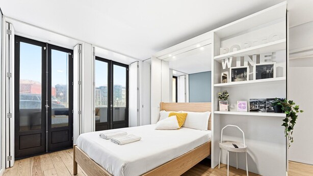 A modern bedroom with a bed, a white chair, a bookshelf, and a city view.