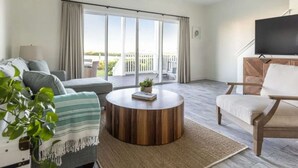 A modern living room with a sofa, a round coffee table, a TV, and a balcony with curtains. - Paddleboarding Paradise | Nature Center. Pool. Four Bedroom Townhome (Duck Key)