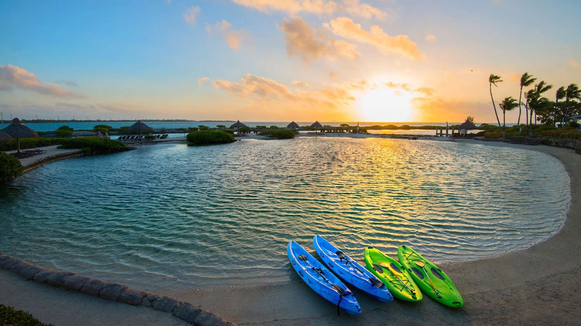 A serene waterfront resort with a curved shoreline, kayaks, and thatched-roof structures.