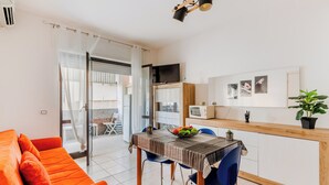 Ein helles Wohnzimmer mit einer Couch, einem Esstisch mit Stühlen, einer Küchenzeile mit einer Mikrowelle und einem Balkon mit Blick.