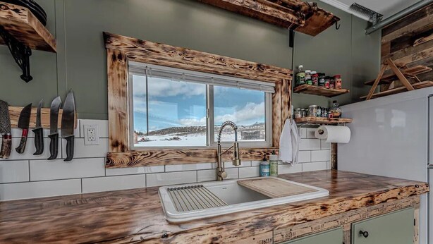 A kitchen with a rustic wooden countertop, a built-in sink, and a window with a snowy landscape view. - Farm Stay, 1-bedroom custom built shipping container on the river. (Ovando)