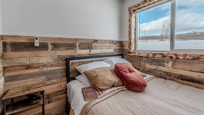 A bedroom with a wooden headboard, a bed with pillows, a book, and a window with a snowy landscape view. - Farm Stay, 1-bedroom custom built shipping container on the river. (Ovando)