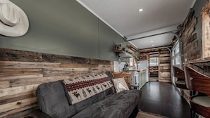 A cozy living area with a sofa, a hat on the wall, and a rustic wooden wall. - Farm Stay, 1-bedroom custom built shipping container on the river. (Ovando)