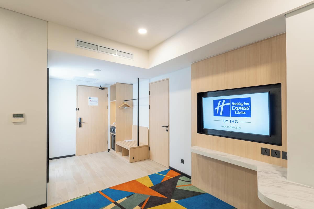 Een hotel lobby met een receptiebalie, een televisiescherm waarop de naam van het hotel staat, en een kleurrijk geometrisch vloerkleed.