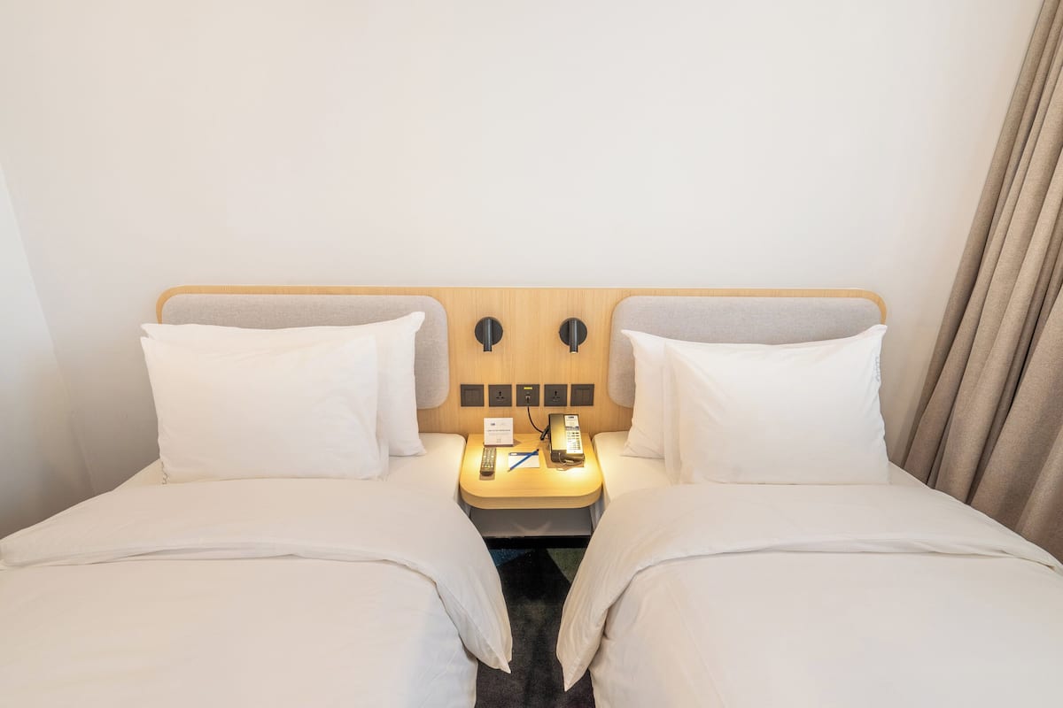 Een hotelkamer met twee bedden, een houten hoofdbord, een nachtkastje met een telefoon en notitieboekje, en een gordijn aan de rechterkant.