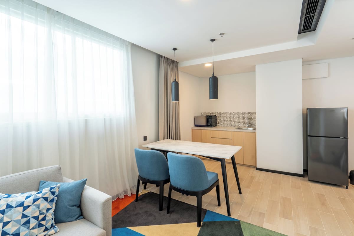 Een gezellige, compacte leefruimte met een bank, twee stoelen, een eettafel, een keuken met een magnetron en koelkast, en een raam met dunne gordijnen.