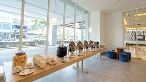Een buffetopstelling met diverse voedselcontainers en bestek op een lange tafel, grote ramen en een modern interieur.
