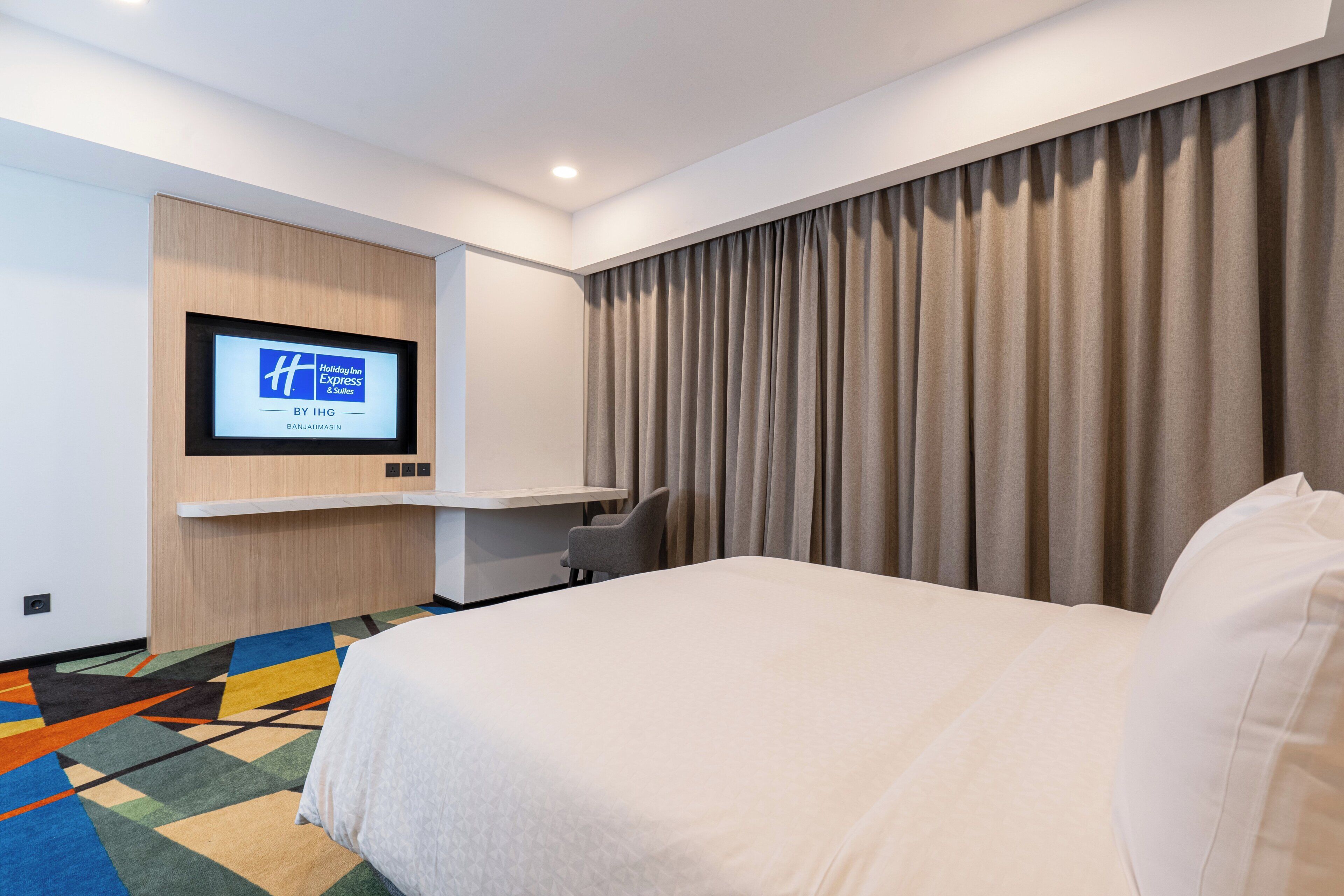 Une chambre d’hôtel avec un lit, un téléviseur à écran plat fixé au mur, un bureau et une moquette colorée.