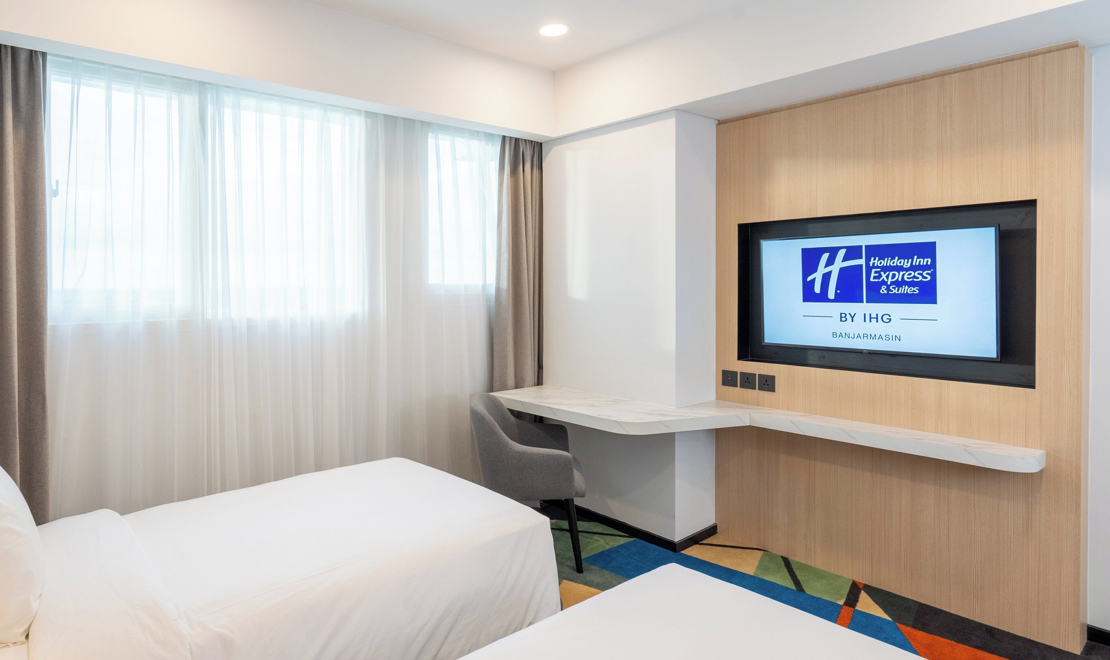Habitación de hotel con cama, escritorio con silla, televisor montado en la pared y ventana con cortinas.