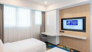 Habitación de hotel con cama, escritorio con silla, televisor montado en la pared y ventana con cortinas.