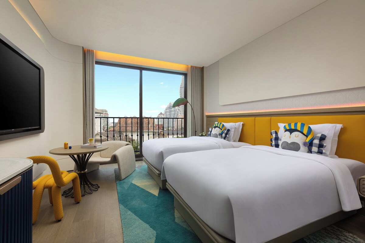 A hotel room with two beds, a TV, a small table, and a balcony with a view of the city.