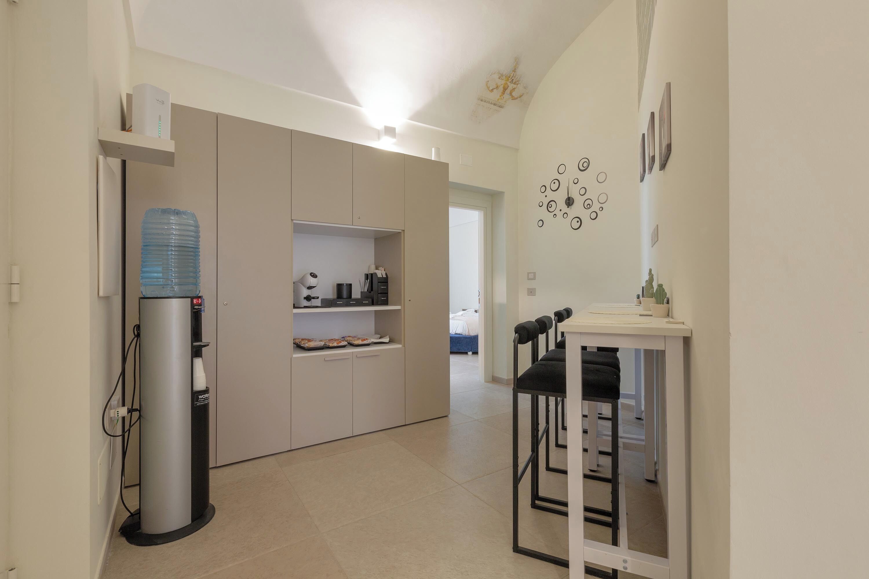 Eine moderne Küche mit einem integrierten Kühlschrank, Wasserspender und Kaffeemaschine. Ein kleiner Essbereich mit Tisch und Stühlen ist ebenfalls vorhanden.