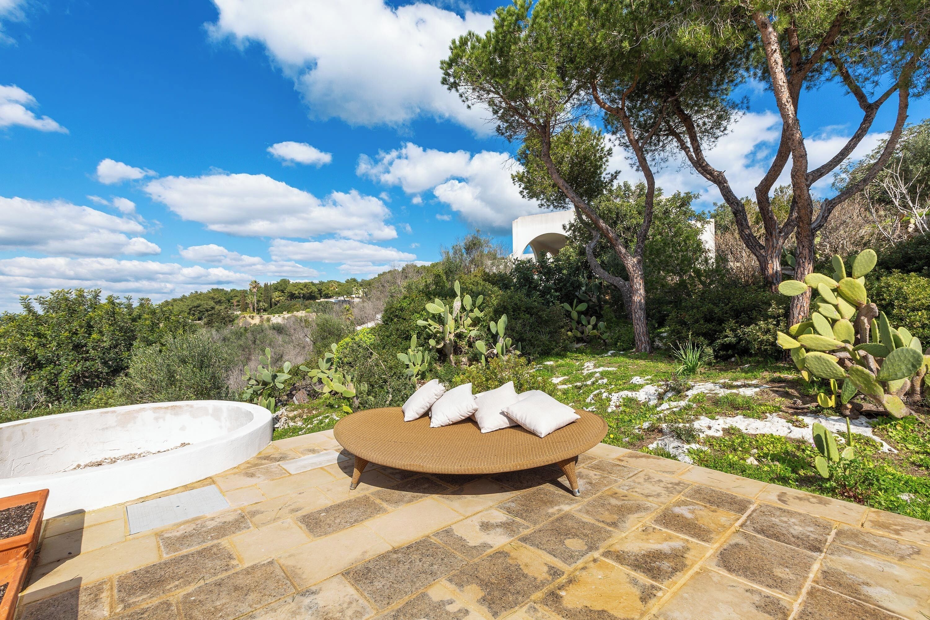 Un área de estar al aire libre con un sofá de mimbre redondo y cojines, una piscina de piedra y follaje circundante.