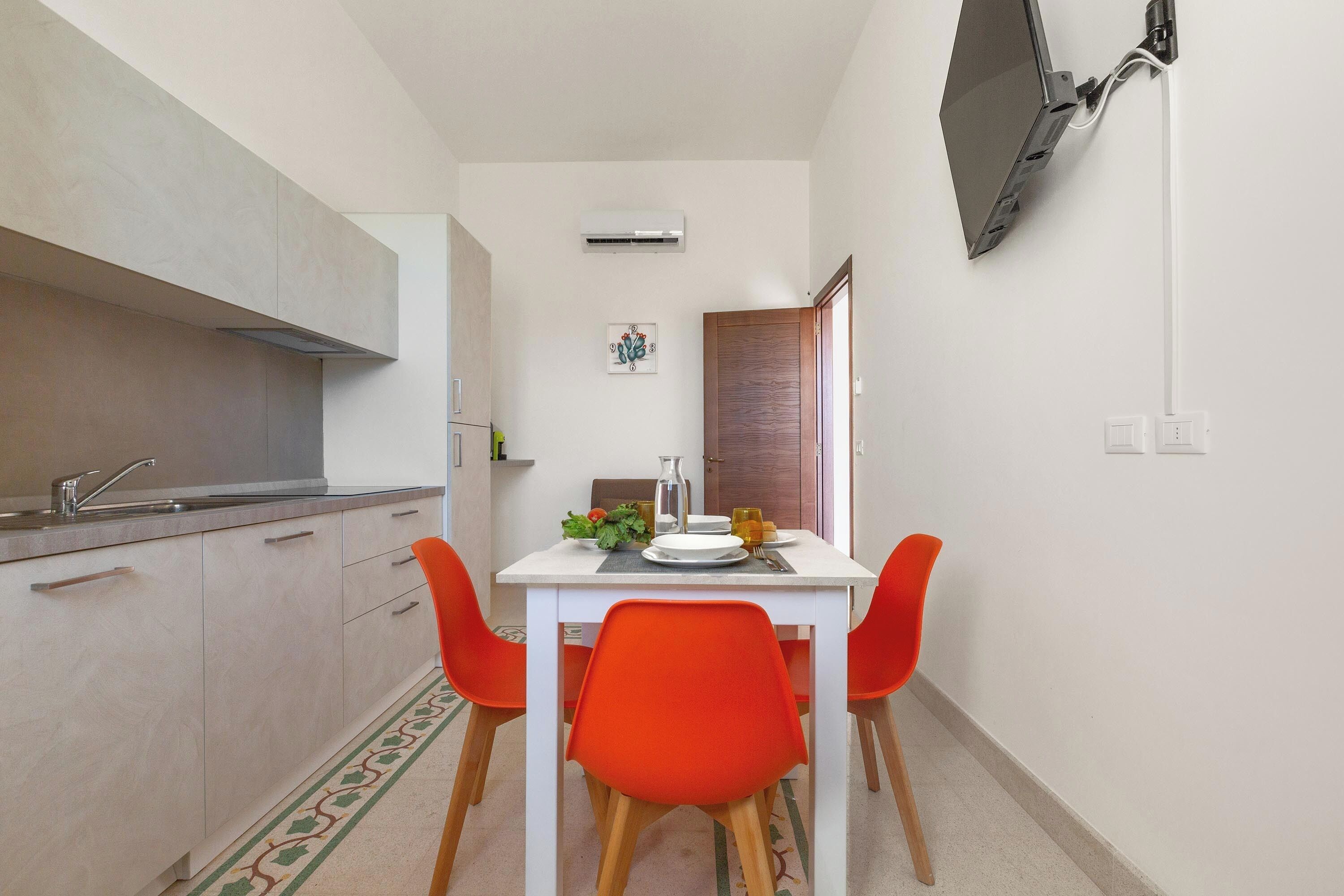 Une cuisine compacte avec un coin repas, un téléviseur à écran plat fixé au mur et une petite table accompagnée de deux chaises orange.
