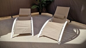 Twee beige loungestoelen met witte frames, geplaatst op een betegeld terras met een bruine achtergrond.