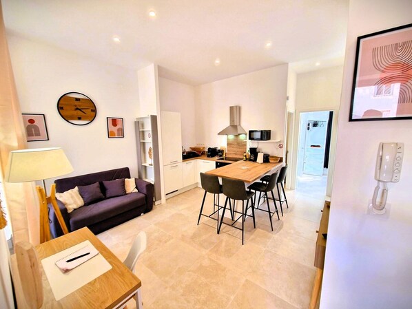 A modern living room with a sofa, a dining table with chairs, a kitchen area with a microwave, and a phone on the wall.