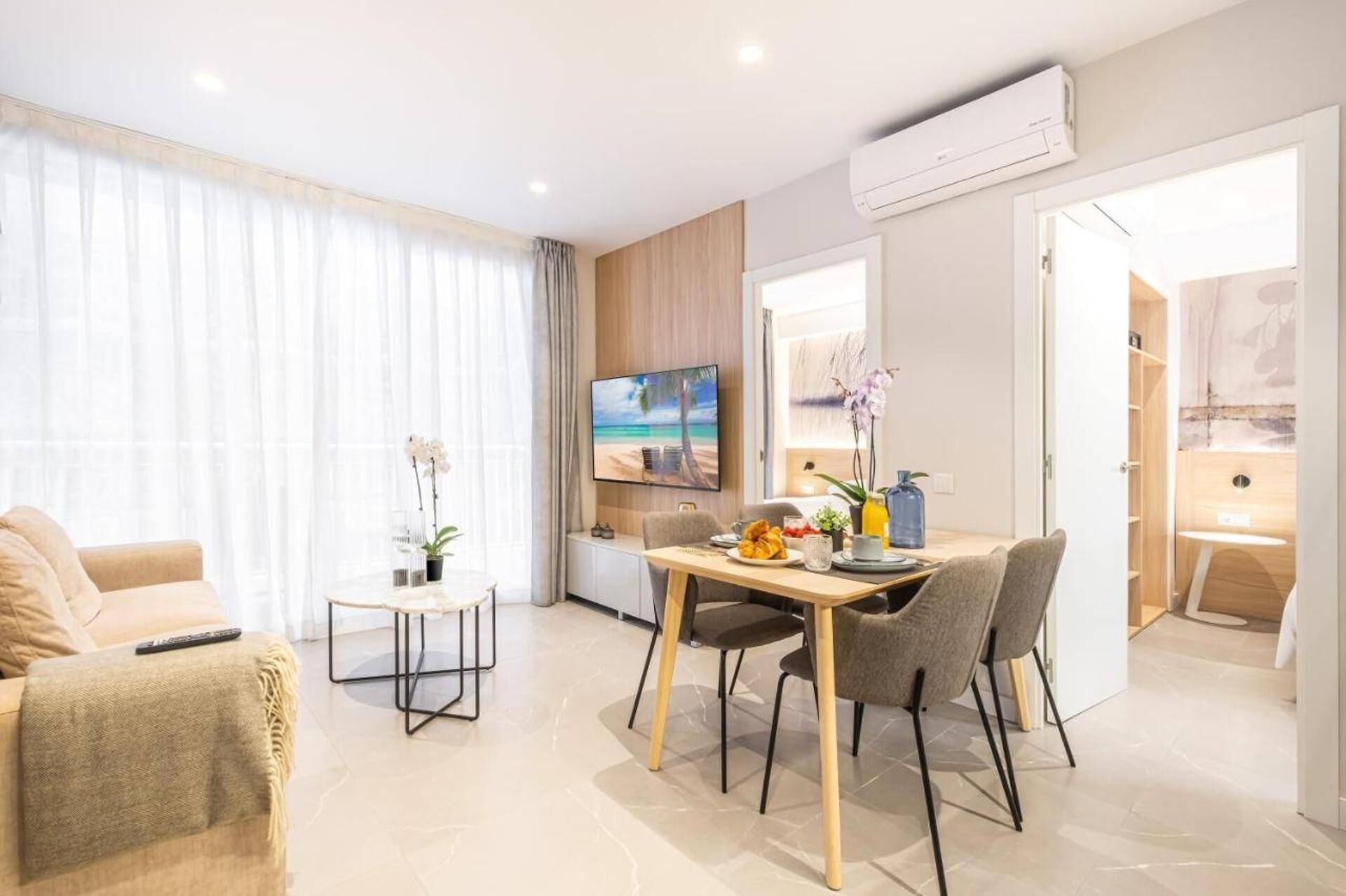 A modern living room with a dining area, a TV mounted on the wall, and a view of the beach through the window.
