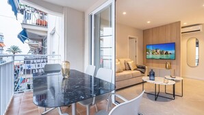 Een moderne woonkamer met een glazen eettafel, witte stoelen, een bank en een televisie aan de muur.