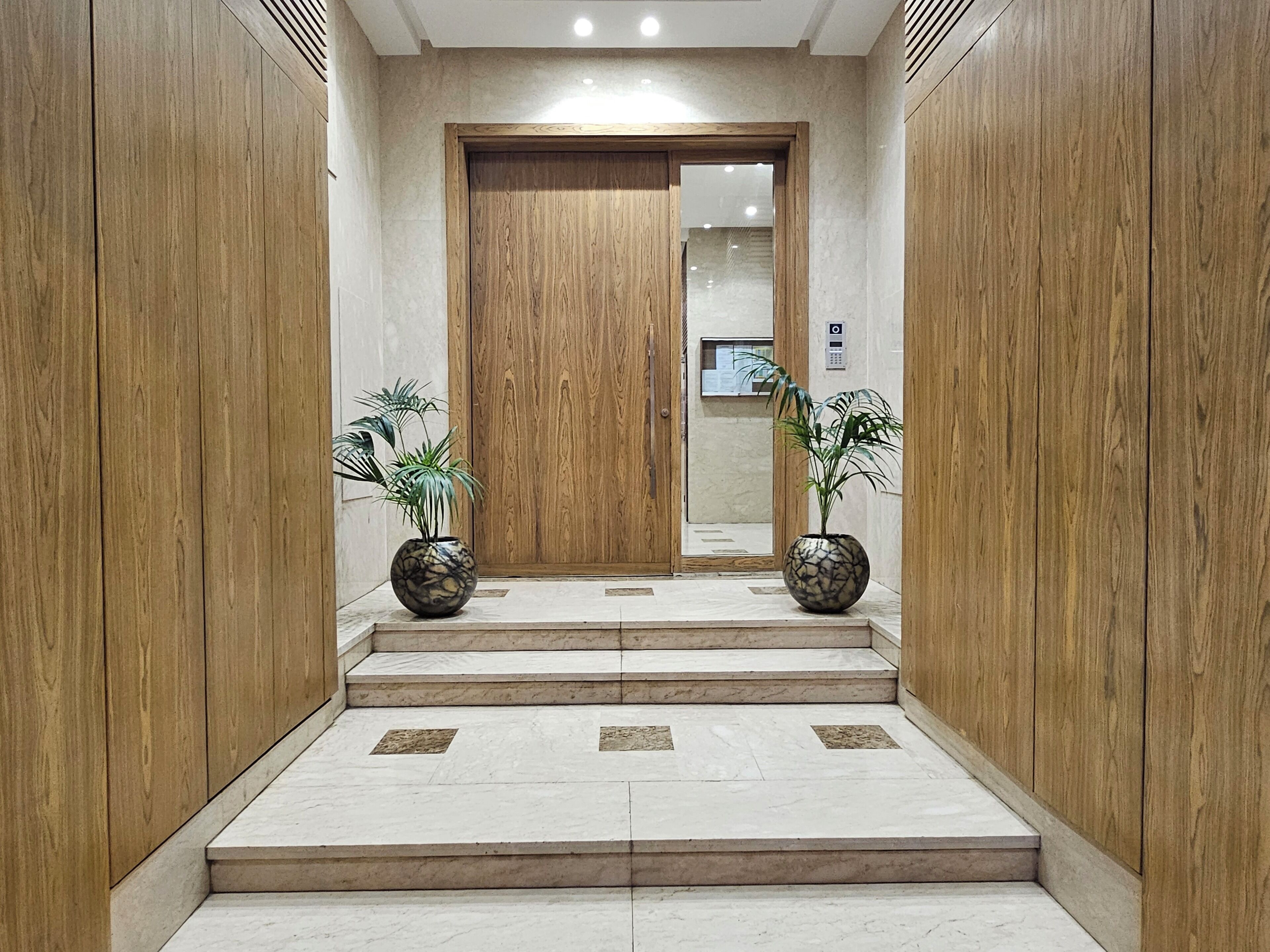 Un hall d’hôtel moderne avec une porte en bois, des marches en marbre et deux plantes en pot.