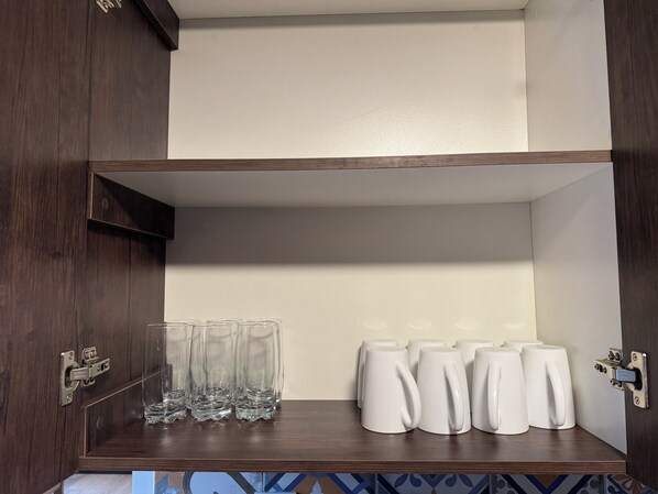 Un mueble de madera con un estante que contiene vasos de vidrio transparentes y tazas blancas.