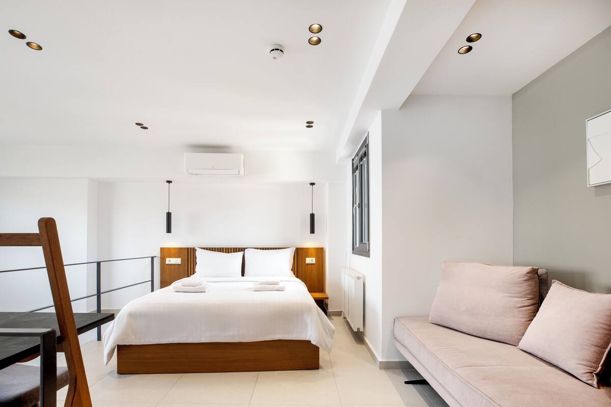 A modern hotel room with a bed, bedside tables, a sofa, and a window.