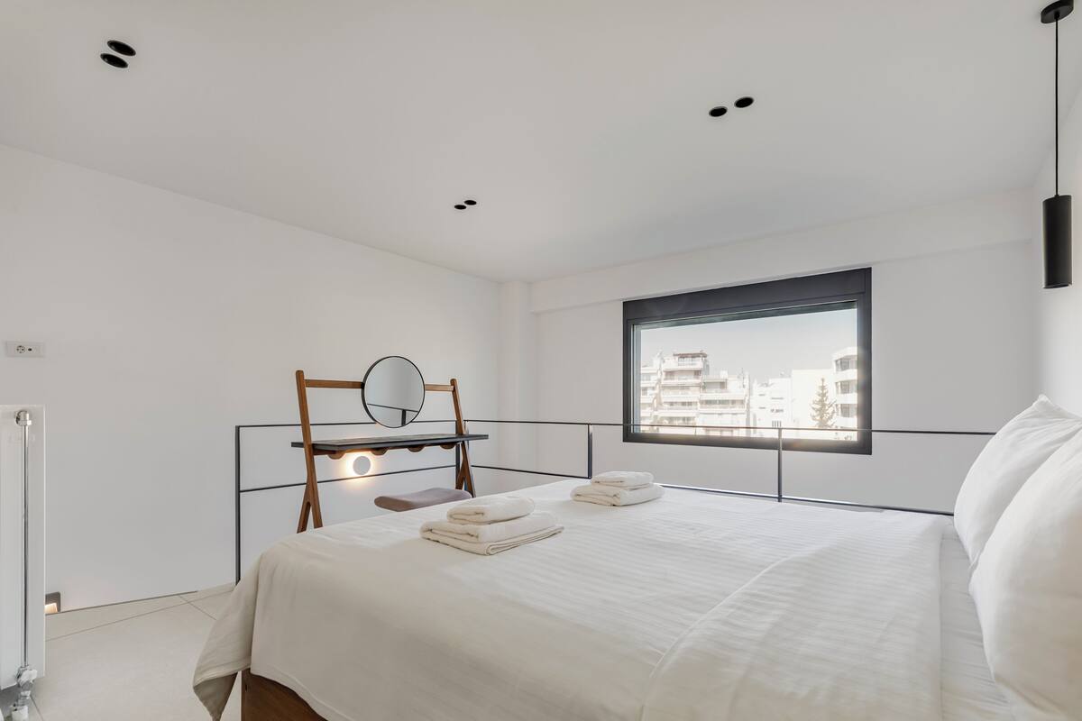 A modern hotel room with a large bed, a bedside table with a mirror, a window with a view of buildings, and a ceiling with recessed lighting.