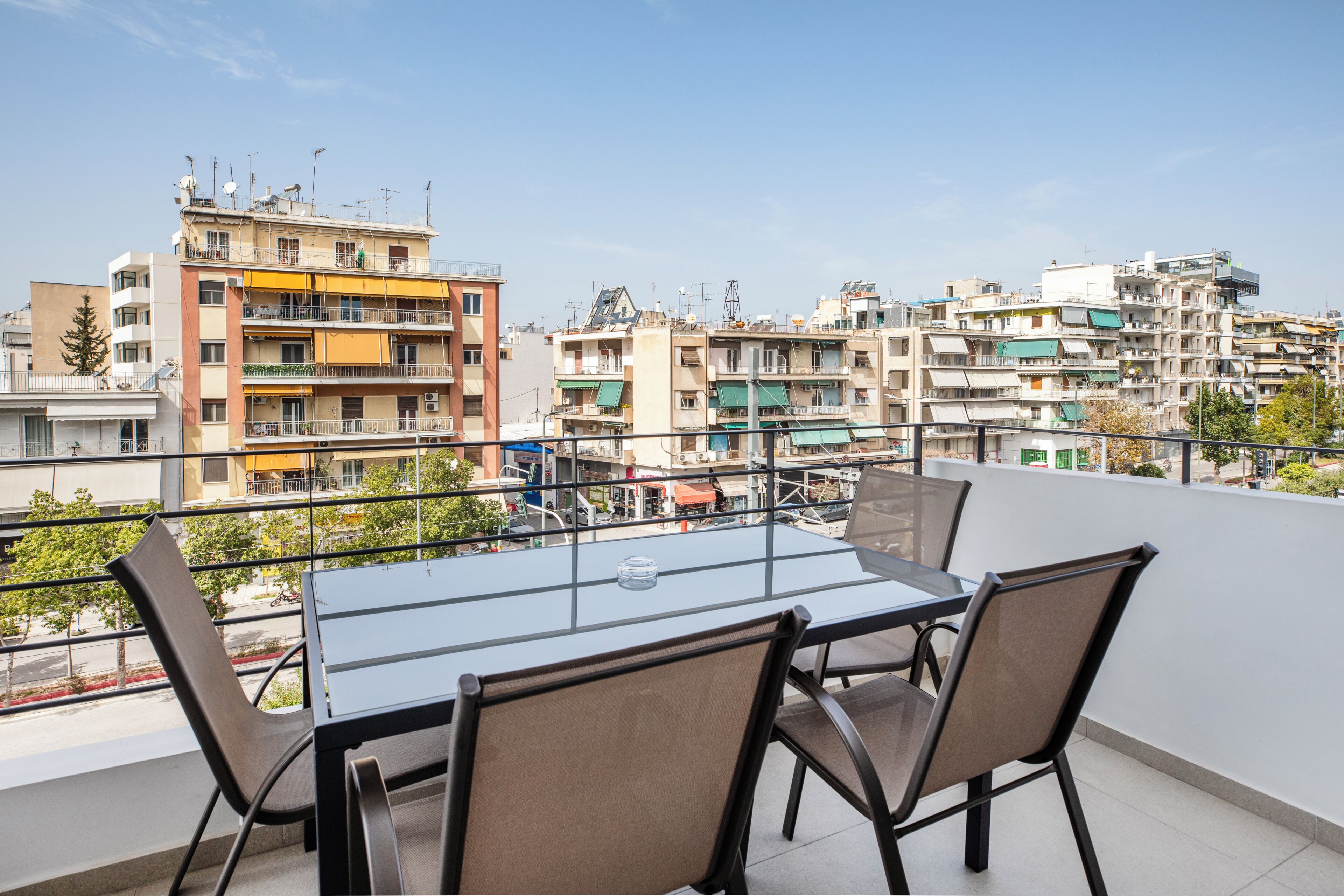 A balcony with a glass table and chairs, overlooking a densely packed urban area with multi-story buildings.