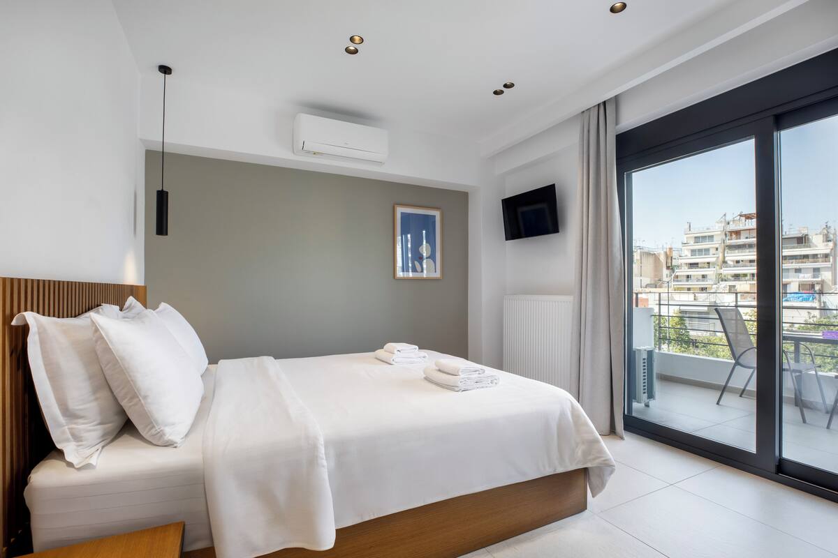 A modern hotel room with a bed, a flat-screen TV, a window with a city view, and a balcony.