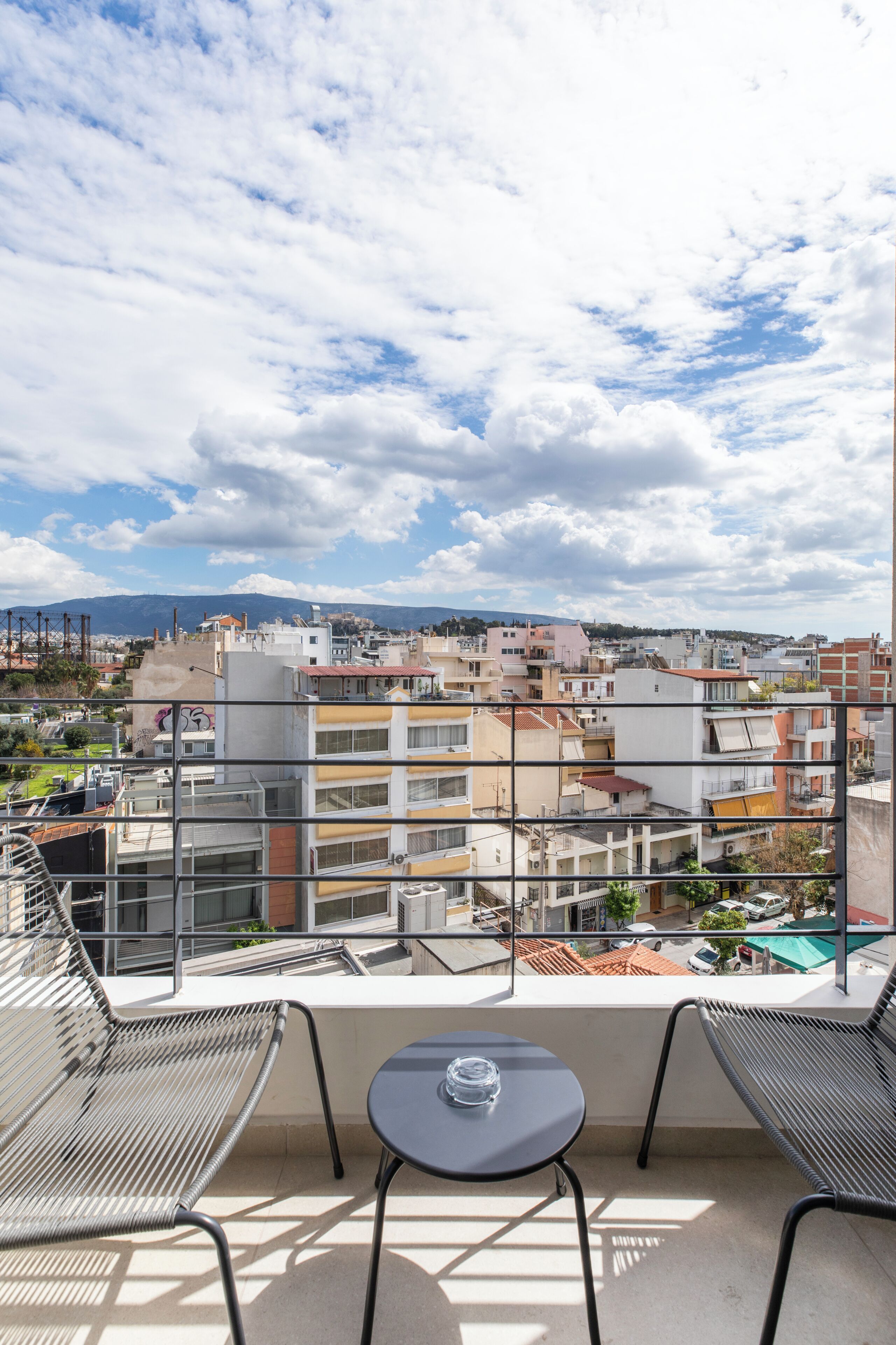 A balcony with a table and chairs, overlooking a cityscape with buildings and a distant mountain.