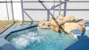 A hot tub with a rocky waterfall feature, surrounded by a wooden deck and a white wall. - 4 Miles to Disney! Private pool -5 Bedroom Home (Kissimmee)
