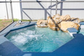 A hot tub with a rocky waterfall feature, surrounded by a wooden deck and a white wall.