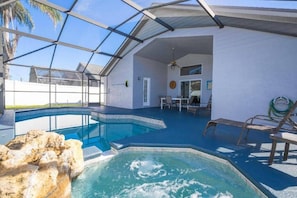 A swimming pool with a glass enclosure, a hot tub, and a patio area with chairs and a table.