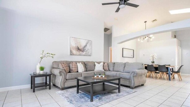 A living room with a sectional sofa, a coffee table, a side table with a plant, a painting on the wall, and a dining area in the background. - 4 Miles to Disney! Private pool -5 Bedroom Home (Kissimmee)