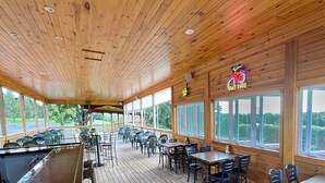A covered outdoor seating area with wooden tables and chairs, large windows, and a view of the outdoors. - Golf, Outdoor Bar, Restaurant - The Forest Haven (Tully)