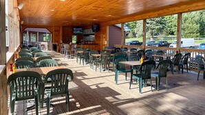 A covered outdoor dining area with wooden tables and chairs, a bar area, and large windows overlooking a parking lot. - Golf, Outdoor Bar, Restaurant - The Forest Haven (Tully)