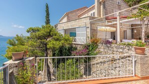 A coastal villa with a stone facade, a terrace with potted plants, and a view of the sea.