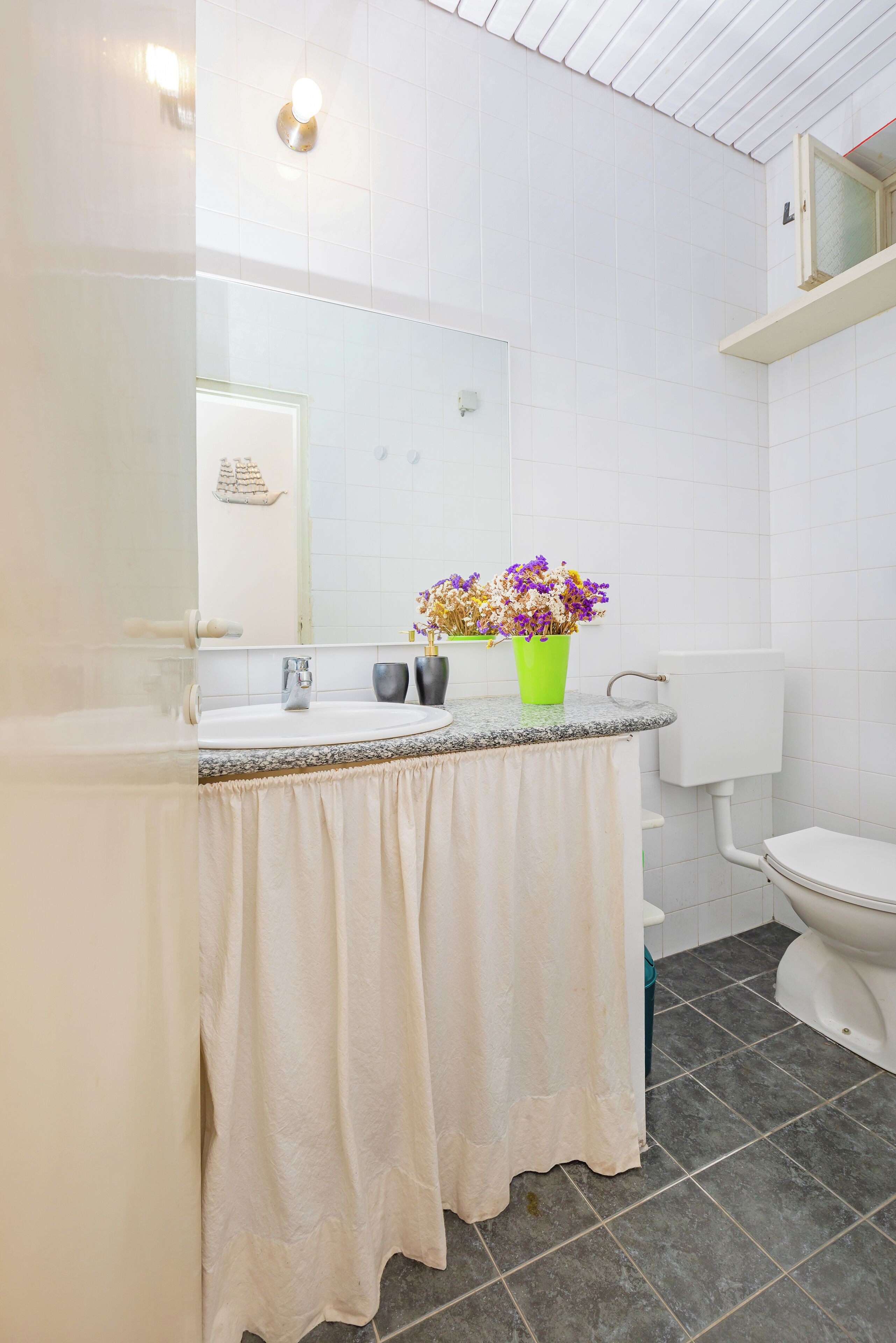 Un baño con lavamanos blanco, un espejo, un inodoro y un jarrón verde con flores.