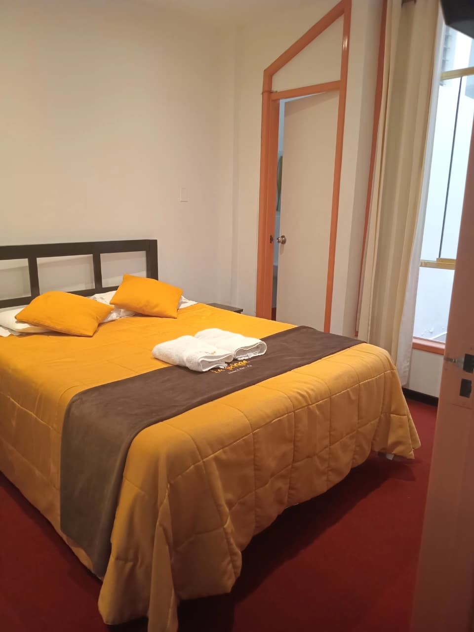 A hotel room with a bed, a wooden headboard, a yellow bedspread, and a red carpet.