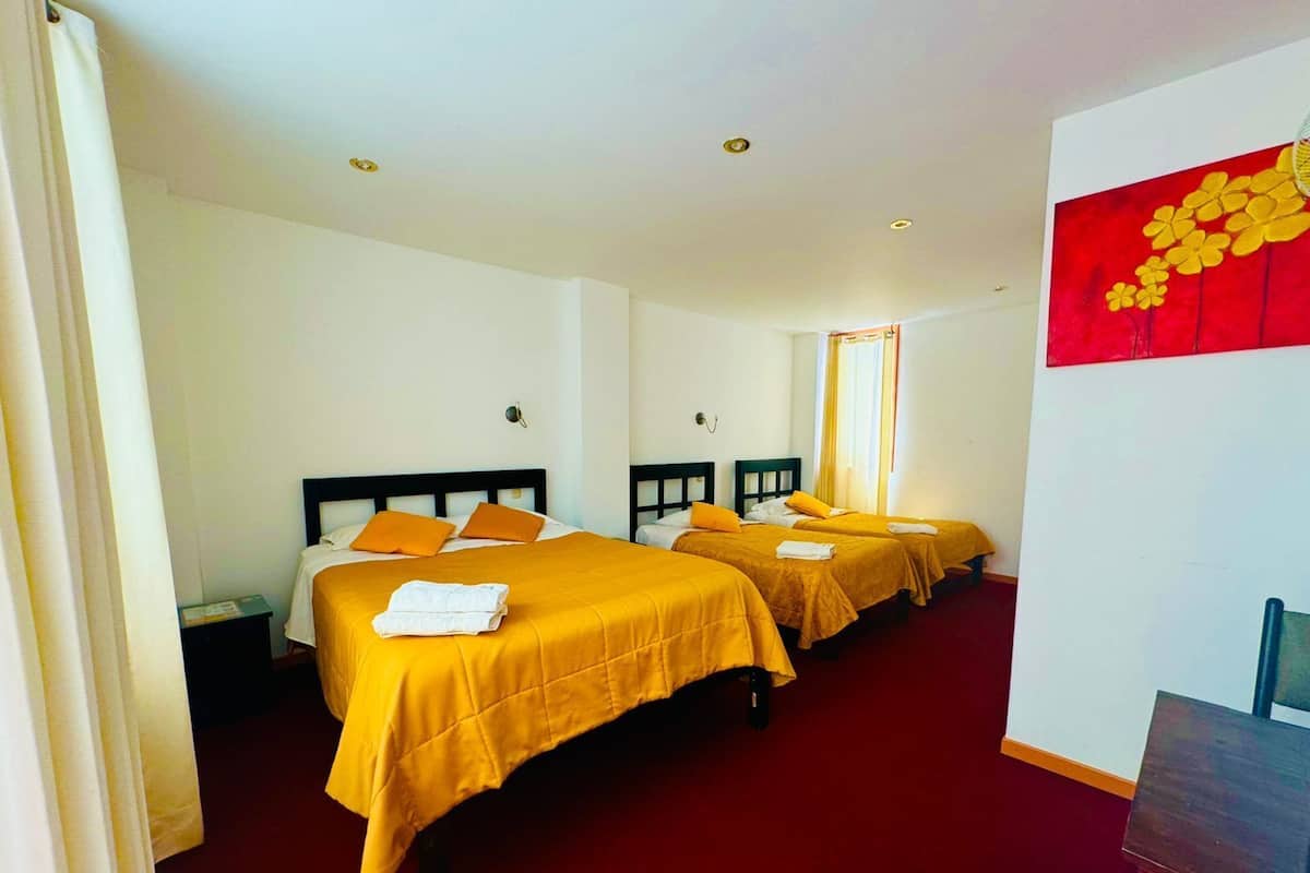 Habitación de hotel con dos camas, cada una con una colcha amarilla y almohadas anaranjadas. Hay una alfombra roja en el suelo, y un cuadro de flores amarillas en la pared.
