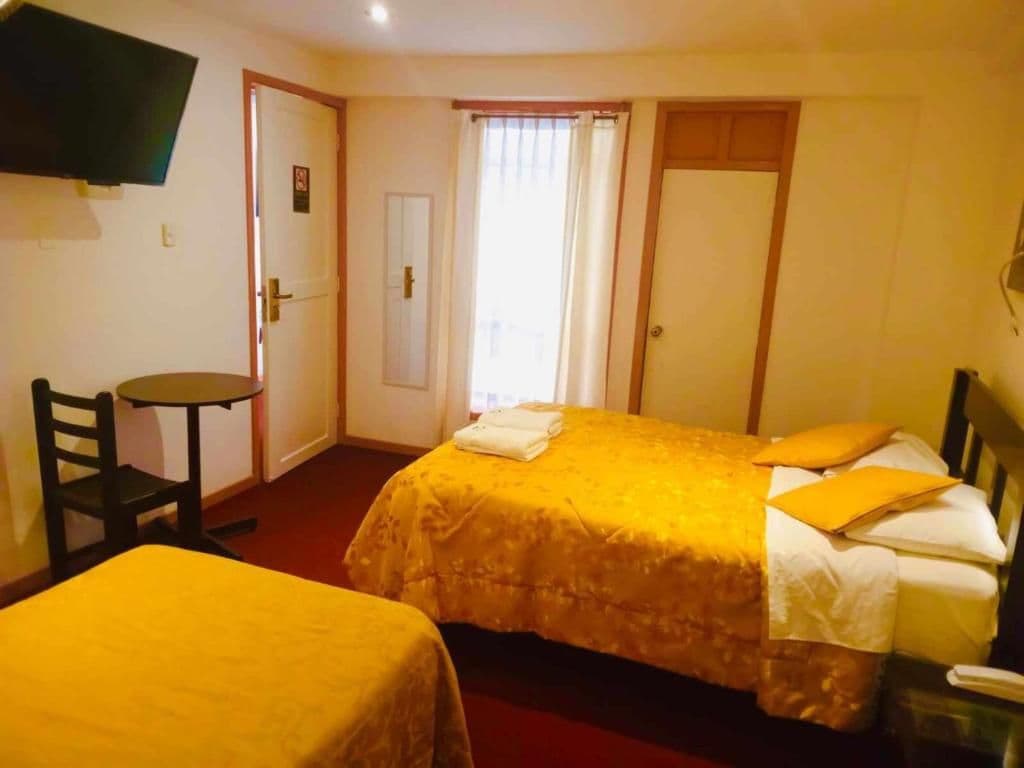 Habitación de hotel con dos camas, televisión montada en la pared, una mesita y puerta que da a un balcón.