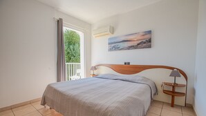 Un dormitorio con cama, mesita de noche, lámpara, un cuadro y vistas a zonas verdes a través de una ventana.