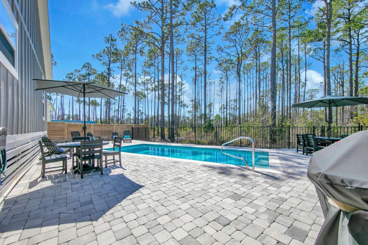 A pool area with a clear view of the pool, surrounded by tall trees and a paved patio with outdoor furniture.