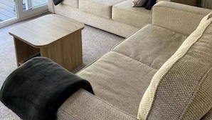 A living room with a beige sofa, a wooden coffee table, and a black throw blanket. There are curtains on the windows, and a sliding glass door leads to an outdoor area.