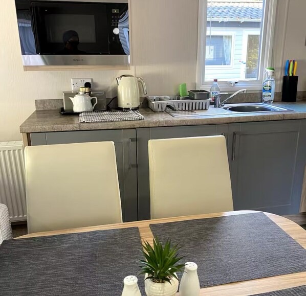 Una cocina compacta con microondas, tetera y lavabo. Hay una mesa de comedor con sillas y una pequeña planta en maceta.