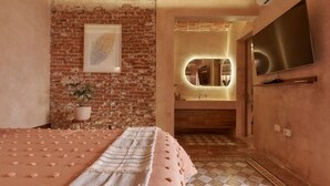 Un dormitorio con pared de ladrillo, cama con colcha rosa, planta en maceta y un espejo grande sobre el baño.