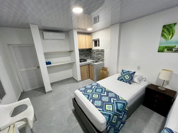 A compact hotel room with a bed, a small kitchenette, a bathroom, and a seating area.