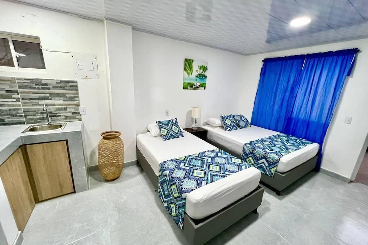 A modern hotel room with two beds, a small kitchenette, a decorative vase, and a blue curtain.